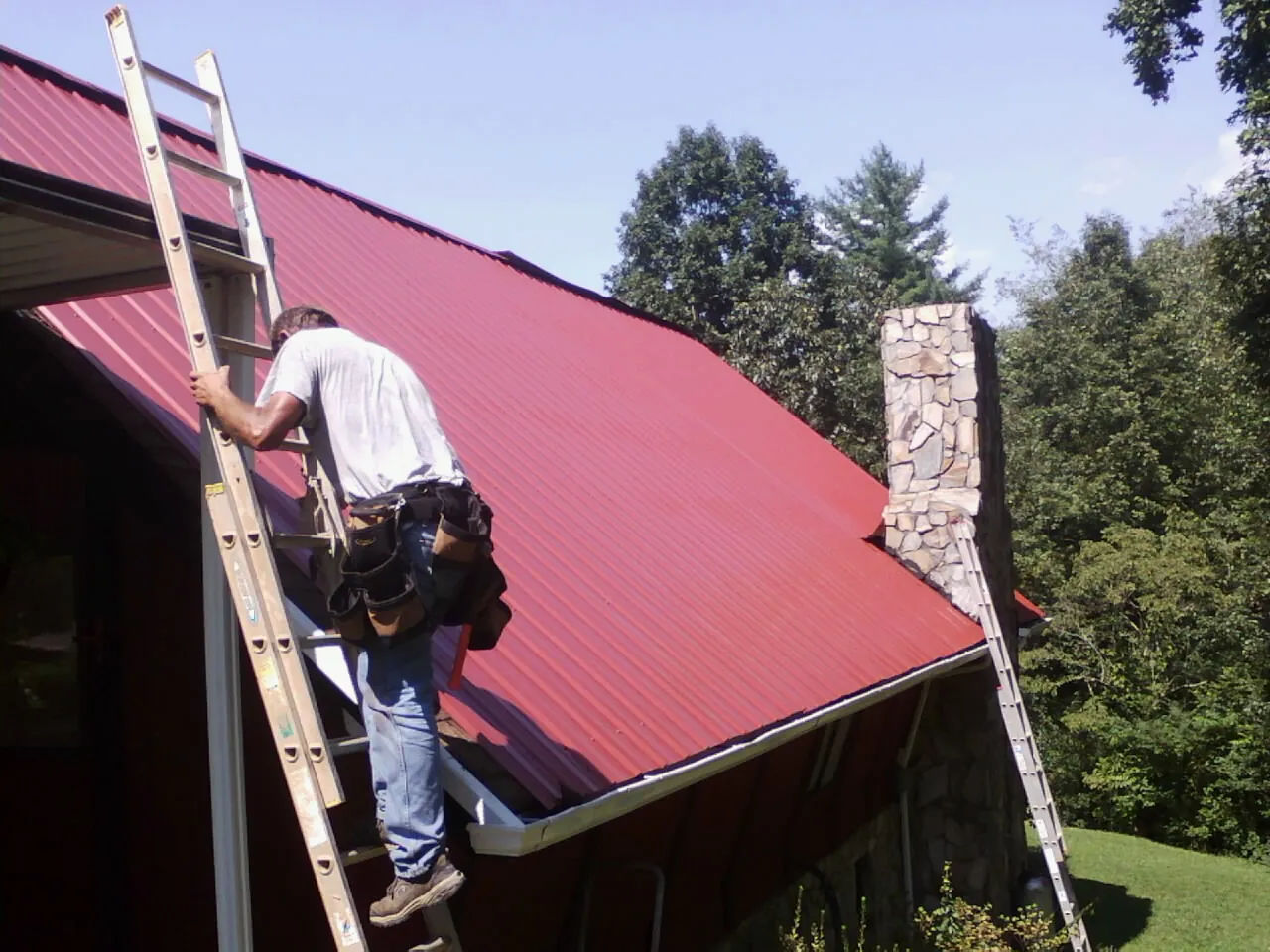 Roofer on a ladder beside a red metal roof and stone chimney — Soffit & Fascia Repair by Roofing Brothers in Wakarusa